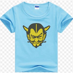 Puede incluir: Camiseta azul claro con un diseño de cara de diablo amarillo y negro. El diseño muestra un diablo con cuernos y perilla. La camiseta tiene mangas cortas y cuello redondo.