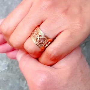 May include: A gold ring with a detailed design featuring a central flower motif. The ring is worn on the finger of a person's hand.