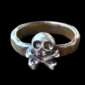 Anillo con calavera y tibias cruzadas, en bronce o plata, a elegir el tamaño y el metal. Indícame el tamaño y el metal que prefieres.