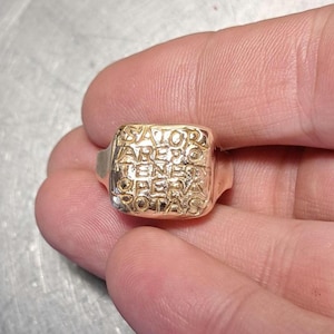Könnte beinhalten: Ein goldfarbener Siegelring mit quadratischer Oberfläche, in die das Sator-Quadrat als lateinisches Palindrom eingraviert ist. Die Wörter SATOR, AREPO, TENET, OPERA und ROTAS sind in einem fünf mal fünf Raster auf dem Metall eingeprägt.
