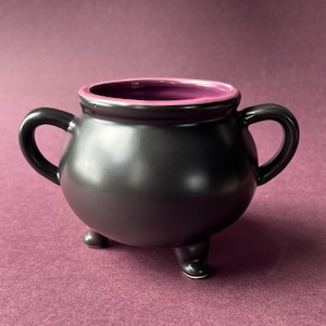 May include: A matte black cauldron-shaped bowl with two side handles and three small feet. The interior of the bowl is a vibrant purple color, contrasting with the dark exterior. The vessel has a rounded body and a wide, open top rim.