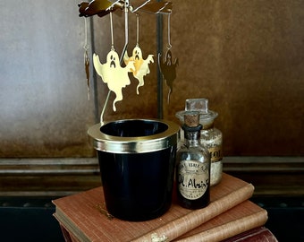 May include: A black candle holder with a gold rim and a gold-coloured mobile featuring ghost figures. The candle holder sits atop a stack of antique books. A small bottle with a label is also visible.