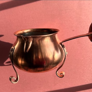May include: A polished copper pot with a rounded body, a wide circular opening, and a metallic interior. The pot features three curved metal legs attached with rivets and a single flat handle extending from the side, showcasing a warm metallic finish.