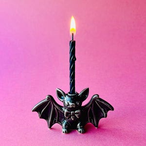 Peut inclure: Un bougeoir noir en forme de chauve-souris avec une bougie noire allumée. Le bougeoir a des ailes détaillées et un nœud papillon. Le fond est de couleur rose uni.