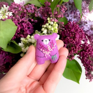 May include: A small, purple felt bear holding a bouquet of lavender. The bear has embroidered details, including a white face and black eyes. The background features a vibrant arrangement of purple and white lilacs.