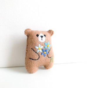 May include: A brown felt bear holding a bouquet of blue and white embroidered flowers with yellow centres.