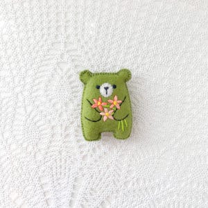 以下が含まれることがあります： ピンクと黄色の花束を持った、手作りの緑色のフェルトのクマ。クマは白い鼻と黒いステッチのディテールがあります。花は黄色の中心と緑色の茎を持っています。クマの高さは約8cmです。