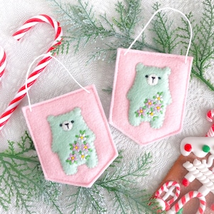 May include: Two pink felt pennants with a light green bear design and floral accents. Each pennant has a white string for hanging. The background includes candy canes, greenery, and a gingerbread house, suggesting a holiday theme.