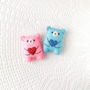 May include: Two felt bear ornaments, one pink and one blue, each holding a glitter heart. The pink bear has a red heart, and the blue bear has a blue heart. Both have black stitched details and white accents.