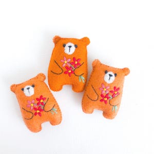 May include: Three orange felt bear ornaments with embroidered details. Each bear has a white face, black eyes, and a bouquet of pink and red flowers. The bears are approximately 3 inches tall.