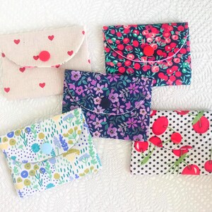 May include: Five fabric pouches with flap closures and button accents. The pouches feature various floral and heart patterns in shades of red, blue, purple, and green. The pouches are arranged on a white textured surface.