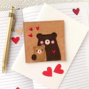 May include: A tan leather square with two felt bear figures, one brown and one tan, with pink cheeks and red heart accents. Red heart-shaped stickers and a gold pen are also visible on a lined notebook page.