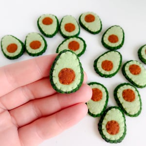 May include: A collection of felt avocado slices. Each slice features a light green exterior, a dark green rind, and a brown seed. One avocado slice is held in a hand, with several others scattered around.
