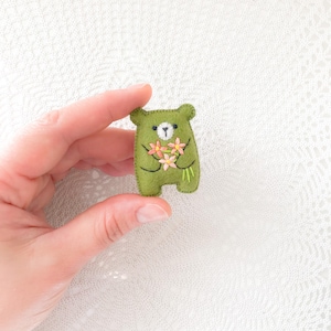 以下が含まれることがあります： 手作りのオリーブグリーンのフェルトベアブローチ。ピンクと黄色の刺繍の花束を持っています。クマは白い顔と黒い目です。魅力的なアクセサリーです。