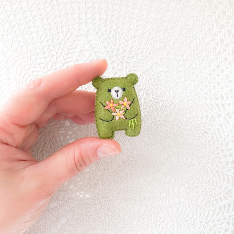 以下が含まれることがあります： 手作りのオリーブグリーンのフェルトベアブローチ。ピンクと黄色の刺繍の花束を持っています。クマは白い顔と黒い目です。魅力的なアクセサリーです。