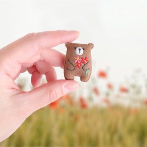 May include: A small brown felt bear holding a bouquet of red and pink flowers. The bear is embroidered with a face and has a brown nose.