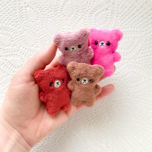 Könnte beinhalten: Vier kleine Teddybären aus Plüsch in Rot-, Braun-, Rosa- und Rosatönen. Jeder Bär hat ein gesticktes Gesicht mit schwarzen Augen und einer kleinen Nase. Die Bären werden in einer Hand gehalten.