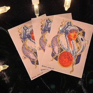 May include: Three stickers featuring a blue and white illustration of a woman with long hair and a fish. The woman is wearing a white headband and a blue and white patterned garment. The fish is a koi fish with orange, white, and black scales. The background is a light pink color. The stickers have the text "@steph_clarks_art" on them.