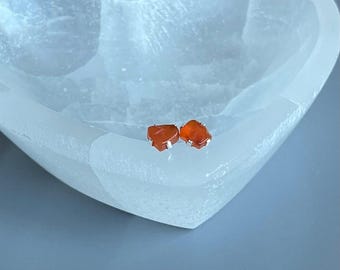 May include: Two small, orange-red carnelian gemstone stud earrings with silver-coloured posts. The earrings are displayed inside a heart-shaped, white, translucent bowl on a grey surface. The earrings are a natural, raw stone shape.