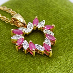 May include: A gold necklace with a pendant featuring a wreath design. The pendant is adorned with oval-shaped pink gemstones and small white gemstones. The necklace rests on a green surface.