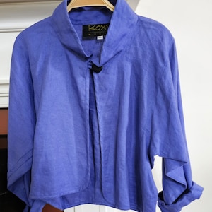 May include: A blue linen jacket with a mandarin collar and a single button closure. The jacket has a relaxed fit and long sleeves. The label on the jacket reads "ROXY" in yellow lettering.