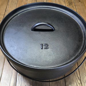 Puede incluir: Una olla holandesa de hierro fundido negro con tapa y asa. La tapa tiene un asa y el número "12" en relieve. La olla tiene un asa de alambre y está sobre una superficie de madera.