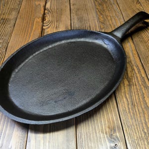 May include: A black, oval cast iron skillet with a handle. The skillet is sitting on a wooden surface with a natural wood grain pattern. The skillet is designed for cooking.