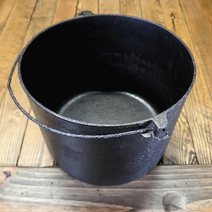 May include: A black cast iron Dutch oven with a handle. The pot is round with a wide opening and a textured surface. The handle is made of the same material as the pot. The Dutch oven is sitting on a wooden surface.