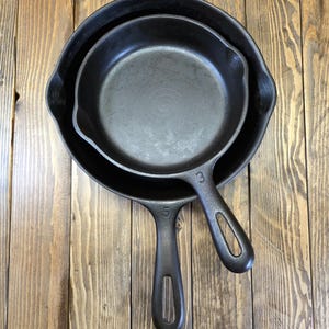 May include: Three nested black cast iron skillets with long handles, sizes 3 and 5, on a wooden surface. The skillets are arranged inside each other, showcasing their different sizes and shapes. The background is a light brown wooden surface.