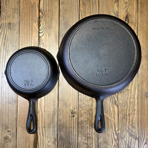 Peut inclure: Deux poêles en fonte noires, l'une marquée "No. 3 6 1/2 IN" et l'autre "No. 7 10 1/2 IN MADE IN USA". Les deux poêles ont une poignée et sont présentées sur une surface en bois. Les poêles sont rondes et ont une finition sombre et mate.