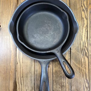 May include: Two nested, well-seasoned cast iron skillets. The larger skillet has a diameter of approximately 10 inches, while the smaller one is about 7 inches. Both have integrated handles and are displayed on a wooden surface.