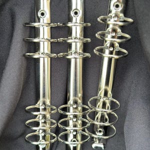 May include: Three silver-colored binder mechanisms with multiple rings, designed for organizing papers. The metal components are arranged vertically against a dark background, with a small collection of additional hardware at the bottom.