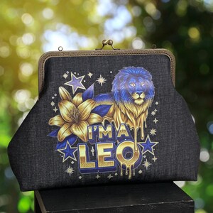 May include: A black denim clutch with a metal clasp. The clutch features a gold and blue lion illustration with the text "I'M A LEO" and floral and star accents. The background is blurred.