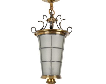 May include: A brass and black metal pendant light fixture with a frosted glass shade. The shade has a grid pattern and is shaped like a cone. The fixture has a decorative scrollwork design.