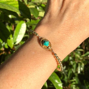 May include: A gold-toned beaded bracelet featuring a central oval-shaped filigree charm set with a single square-cut turquoise stone. The bracelet is constructed from a series of small, uniform metallic beads connected to the central decorative element.