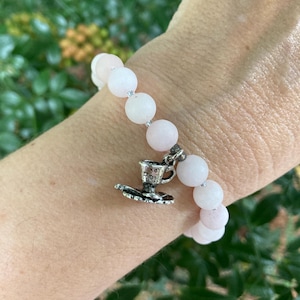 May include: A white beaded bracelet with a silver teacup charm. The beads are round and have a slightly textured surface.