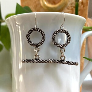 May include: A pair of silver-toned earrings. Each earring features a circular design with a woven texture, connected to a horizontal bar with a similar woven pattern. The earrings are suspended from a simple hook.