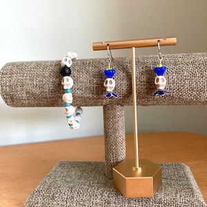 Peut inclure: Un présentoir doré avec deux paires de boucles d'oreilles en forme de crâne. Les boucles d'oreilles sont faites de crânes en céramique blanche et de perles de verre bleues. Un bracelet en céramique blanche en forme de crâne avec des perles turquoise est également présenté sur le présentoir.