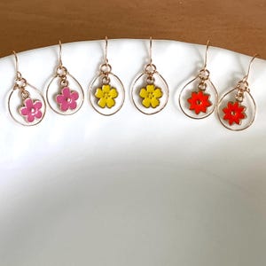 Puede incluir: Seis pendientes colgantes con aros de metal dorado y pequeños dijes de flores. Los dijes están esmaltados en rosa, amarillo y naranja. Cada pendiente tiene un gancho para la oreja, mostrando un diseño simétrico con colores vibrantes.