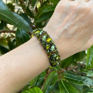 May include: A wide bracelet featuring a dark metal filigree base adorned with numerous olive green and lime green faceted crystals in various teardrop and round shapes, creating a dense, textured, and multi-layered botanical appearance.