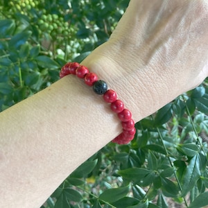 Puede incluir: Un brazalete de cuentas rojas con una cuenta de lava negra. El brazalete se lleva en la muñeca.