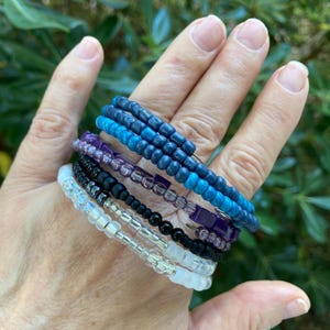 May include: A multi-strand coil bracelet featuring rows of small, round beads in a gradient of colors. The layers consist of matte white, clear, solid black, translucent purple, and two shades of blue beads arranged in a spiral design.