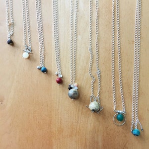 Puede incluir: Ocho collares de cadena plateada dispuestos en fila, cada uno con un colgante único. Los colgantes incluyen varias cuentas esféricas en colores como negro, blanco, azul, rojo, beige y verde, con algunos diseños que incorporan anillos metálicos.