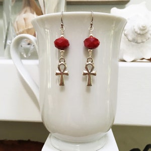 May include: A pair of dangle earrings with red faceted beads and silver ankh charms. The earrings are suspended from silver hooks. The earrings are displayed against a white background.