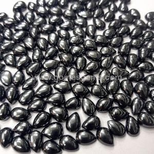 May include: A collection of polished, teardrop-shaped, black gemstones. The stones have a high-gloss finish, reflecting light and creating a dark, lustrous appearance. The gemstones are scattered, showcasing their individual shapes and sizes.