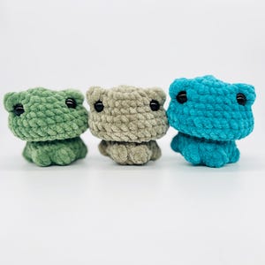May include: Three crocheted frog plushies in green, beige, and blue. Each frog has black eyes and a simple, rounded shape.