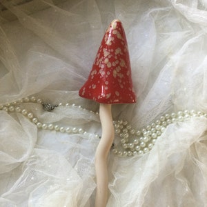 Puede incluir: Una figurita de hongo de cerámica con un sombrero moteado rojo y blanco y un tallo blanco. El hongo está sentado sobre una tela de encaje blanca.