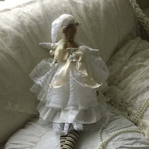 以下が含まれることがあります： 白いレースのドレスを着た白い帽子をかぶった人形。人形は真珠のネックレスをつけた白いクッションに座っています。