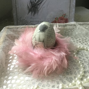 May include: A decorative egg with a vintage image of a woman on it, nestled in a pink feather nest. The egg is sitting on a white lace surface with a pearl necklace.
