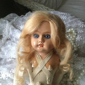 Könnte beinhalten: Eine antike Porzellanpuppe mit blonden Haaren und blauen Augen, die ein weißes Kleid mit einem Kreuzriemen-Detail trägt. Die Puppe liegt auf einem weißen Spitzenstoff mit Perlen.